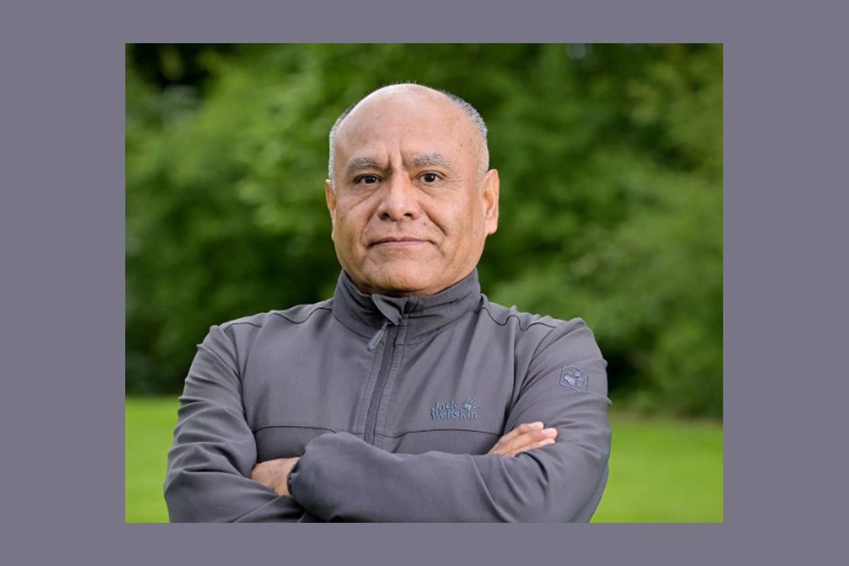 Ein Mann steht mit verschränkten Armen vor einer grünen Wiese. Im Hintergrund sind grüne Bäume verschwommen zu sehen.