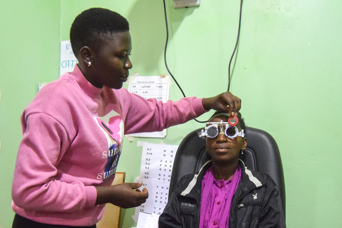 Agnes Nakate (sitzend) und Oliver Nabkiire üben, wie ein Sehtest ausgeführt wird. Beide erhoffen sich von der Ausbildung gute Chancen auf eine Anstellung als Optiker*in.
